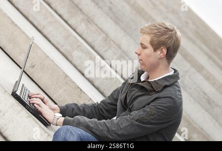 Young blond handsome smiling guy sitting outdoors with laptop computer, Belgium, Europe Stock Photo