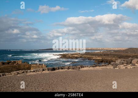 Rocky coast of the Atlantic ocean with small waves. Volcanic rocks of dark brown to black color. Blue sky with white clouds. La Santa, Lanzarote, Cana Stock Photo