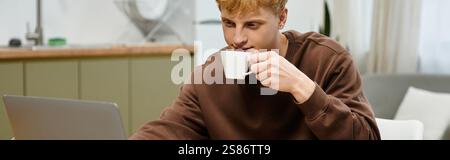 Casual dressed young redhead focuses on his laptop while enjoying a warm drink, banner Stock Photo
