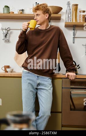 A handsome young man relaxes in his casual outfit, sipping coffee in a cozy kitchen. Stock Photo