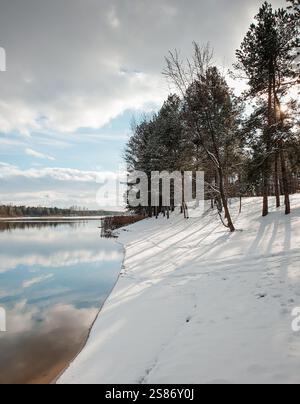 Bank of the river on a sunny winter day. Winter landscape with sun shining through pine forest and cloudy sky. Stock Photo