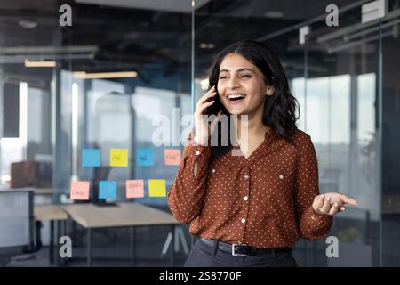 Successful and happy office worker talking on the phone while standing near the window inside the office building. Satisfied businesswoman at workplace communicating with colleagues and clients. Stock Photo