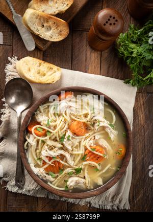 Bowl of chicken noodle soup with carrots on a napkin and baguette slices on a wooden table.  Overhead view Stock Photo