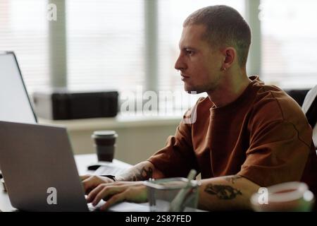 Side view of hard working young male programmer coding on laptop while working at desk in modern IT company office captured against window, copy space Stock Photo