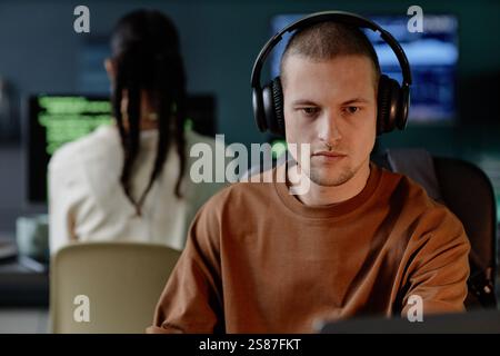 Portrait of senior male developer in headphones intently coding on laptop while working in evening hours in IT company office with colleagues, copy space Stock Photo