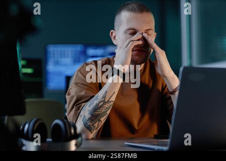 Portrait shot of exhausted from work overload male IT developer sitting with eyes closed at desk working late in office, copy space Stock Photo