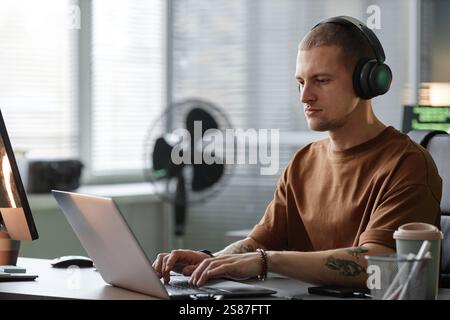 Medium shot of handsome male programmer wearing headphones coding on laptop while working at desk in corporate office of contemporary IT company, copy space Stock Photo