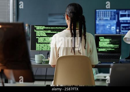 Rear view of Middle Eastern female programmer with braids hairstyle coding on multiple computers at professional workstation in IT company office Stock Photo
