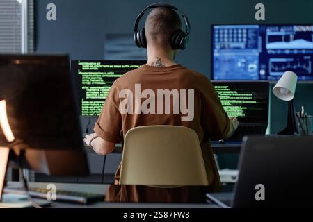 Rear view of male IT developer wearing headphones immersed in writing code on multiple computers at workstation while working alone in office Stock Photo