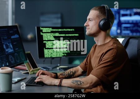 Side view of handsome male programmer wearing headphones working at ease on desktop computer writing code captured against multiple screens in corporate office Stock Photo