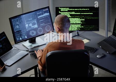 Rear view of male computer programmer at workstation looking at multiple screens with lines of code running while working in office Stock Photo