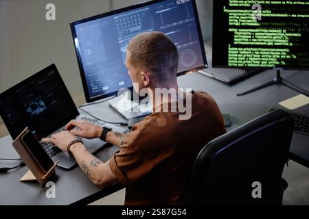 Rear view of junior male programmer typing on laptop keyboard working at desk with multiple computer screens in IT company office Stock Photo