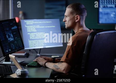 Side view of handsome male IT programmer typing on computer keyboard at workstation writing code in corporate office during evening hours Stock Photo