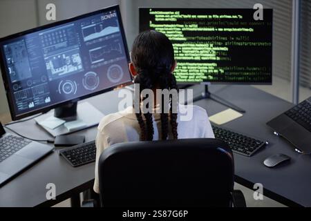 Rear view of female programmer with braids hairstyle coding at desk with several computer screens while working in IT company office Stock Photo