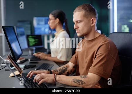Side view of concentrated male IT engineer immersed in writing code on laptop working at large desk with colleague in modern tech company office, copy space Stock Photo