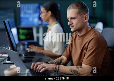 Side view of male IT programmer working at ease typing on laptop while sitting at large desk with colleague in modern tech company office, copy space Stock Photo