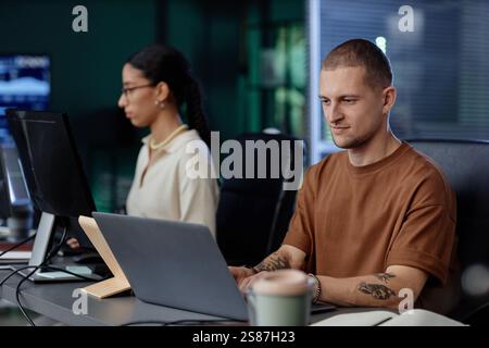 Happy male IT programmer confidently coding on laptop at desk enjoying working in tech company office next to colleagues, copy space Stock Photo