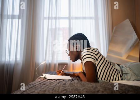 Focused young black woman wearing glasses is lying on her stomach on her bed, diligently writing in a notebook under the warm glow of a small lamp Stock Photo