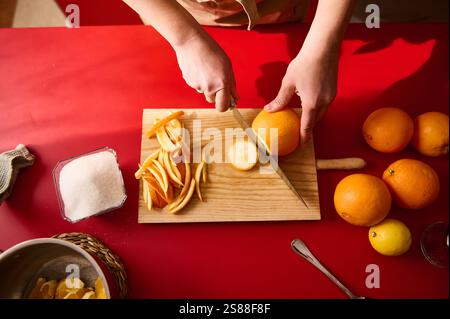 Hands slicing a fresh orange into slices on a wooden board amidst bright kitchen surroundings. Stock Photo
