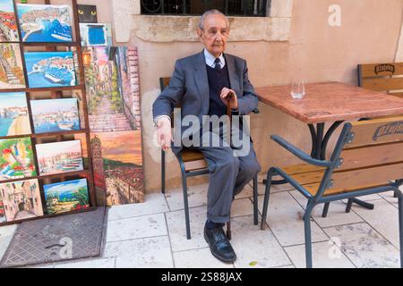 Elderly old senior man at sitting at cafe table with local art paintings painted by local artists, for sale to tourists in the walled Old Town. Dubrovnik, Croatia. (138) Stock Photo