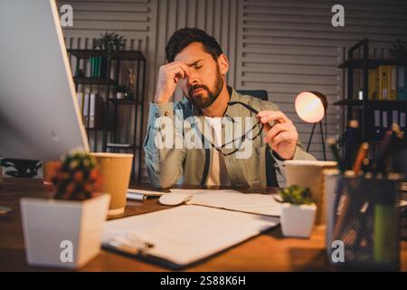 Stressed young professional man working in a busy home office at night, feeling tired and focused on completing tasks under pressure. Stock Photo