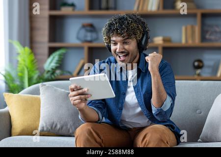 Man sitting with tablet computer at home on sofa, wearing headphones, watching online sports match, celebrating successful results achieved in video game stream. Stock Photo