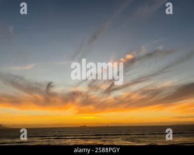 sun going down over the horizon and sea with a ship in the distance and streaky grey clouds in a blue orange sky at sunset in Cape Town, South Africa Stock Photo