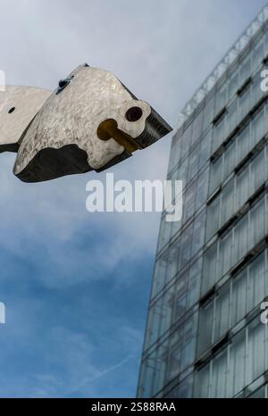 A striking metallic sculpture contrasts a modern glass building, creating an abstract visual interplay against a clear blue sky in an urban setting. Stock Photo