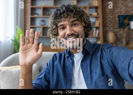 A man looks into the phone camera, happily waves his hand, takes a selfie photo and talks to colleagues and friends remotely using a smartphone app for video chats. Stock Photo
