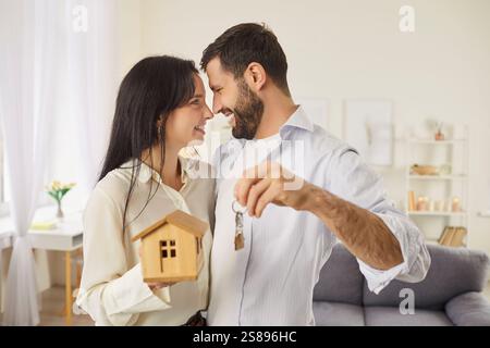 Happy young couple holding keys and toy house, smiling joyfully, celebrating buying their new home. Stock Photo