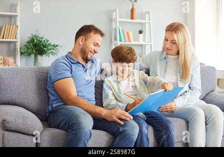 Smiling parents sitting on couch with their son between them, helping him with homework in notebook. Stock Photo