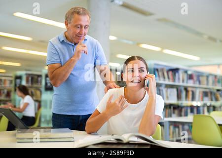 Mature male making silence gesture to happy young female talking on phone in the library Stock Photo