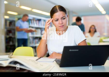 Tired casual female remote worker using laptop and reading books in the library Stock Photo