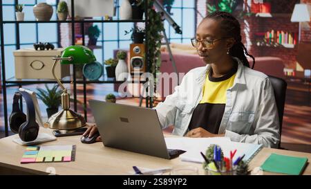 Teleworker using laptop in home office to crosscheck paper documents with online company files. Remote worker typing on notebook, inputting paperwork documentation data, camera A Stock Photo