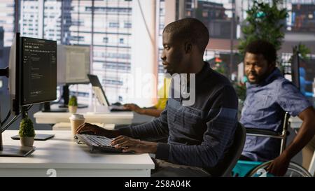 Programmer doing software debugging, asking for help from smiling team leader in wheelchair. Employee fixing coding mistakes, being assisted by cheerful manager with disability, camera A Stock Photo