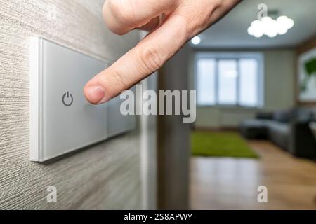 A close-up finger turns on the light on the touch switch. A white modern light switch on a white wall. modern design. Stock Photo