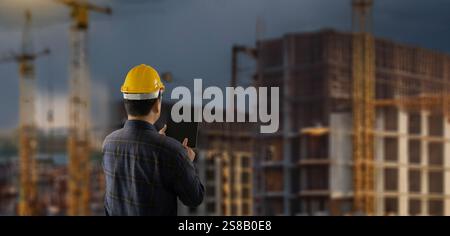 engineer working at site of a large building man wearing a yellow hard hat is looking at a tablet while standing in front of a building. business, Stock Photo