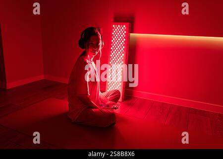 A young woman using a red light therapy panel indoors, wearing headphones Stock Photo