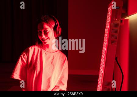 A young woman smiling indoors, wearing headphones, during red light therapy Stock Photo