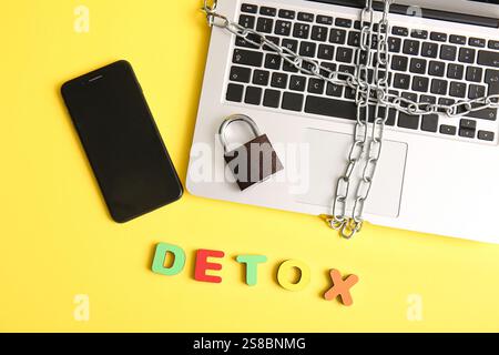Modern laptop with chain, padlock, mobile phone and word DETOX on yellow background Stock Photo