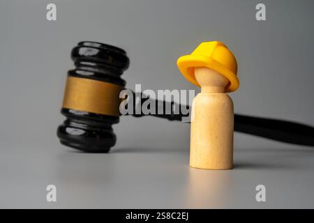 A wooden doll wearing a hard hat stands in front of a gavel. Construction law, worker safety, or legal disputes in the construction industry concept. Stock Photo