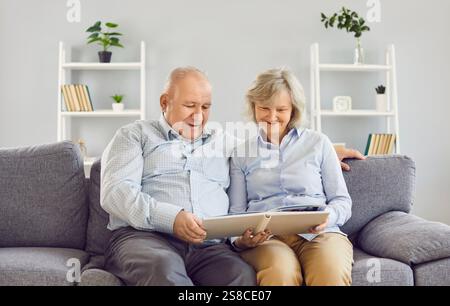 Portrait of happy old gray-haired couple reading a book or watching a photo album on couch at home. Stock Photo
