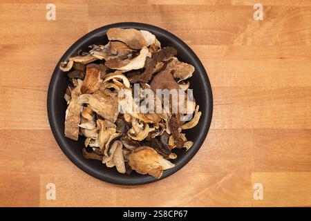 Overhead shot of assorted dried wild mushrooms in a dark ceramic bowl on a natural wood background. Stock Photo