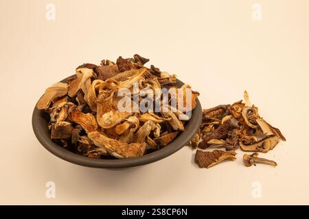 A brown bowl filled with dried mushrooms, with a small pile of mushrooms spilling onto a white background. Stock Photo