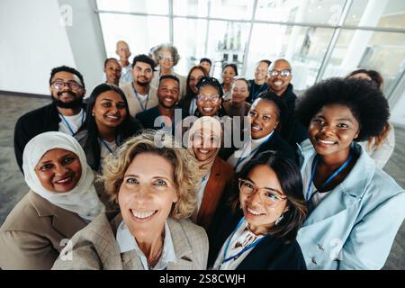 A multiethnic corporate team captured in a group selfie while standing together inside an office building, smiling and showing unity. Stock Photo