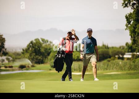 Two sportsmen walking on a golf course, carrying a golf bag, and waving. A perfect depiction of leisure and friendship in a sporting environment. Stock Photo