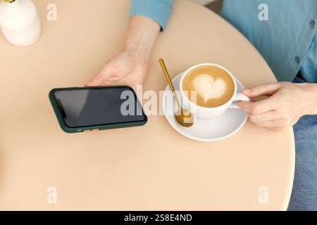 A woman relaxes in a Warsaw cafe, holding a smartphone and a cup of coffee with latte art The cozy setting and modern gadgets create a peaceful atmosp Stock Photo