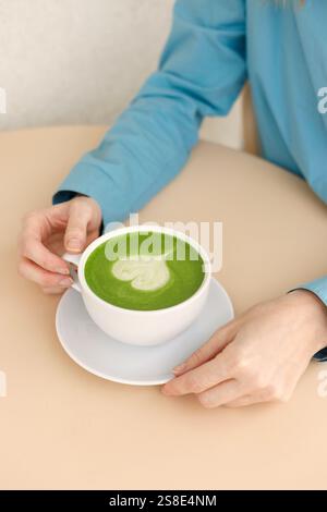 A woman in a blue shirt savors a vibrant green matcha latte with heart-shaped latte art in a cozy Warsaw cafe She gently holds the cup over a minimali Stock Photo