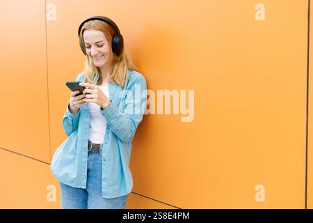 Smiling woman leans against a vibrant orange wall in Warsaw, listening to music on wireless headphones and using her smartphone Casual attire enhances Stock Photo