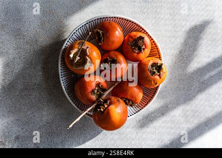 A close-up top view of freshly picked ripe persimmons neatly arranged on a stylish white and blue striped plate. The fruits display a rich orange-redd Stock Photo